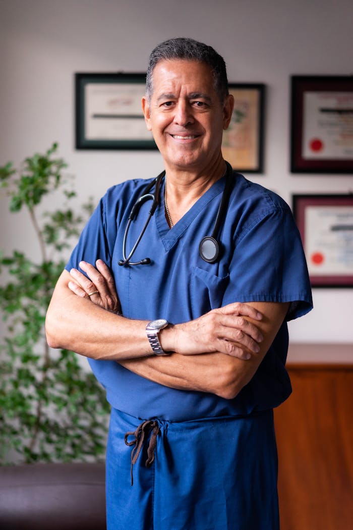 Smiling male healthcare professional in blue scrubs with arms crossed indoors.
